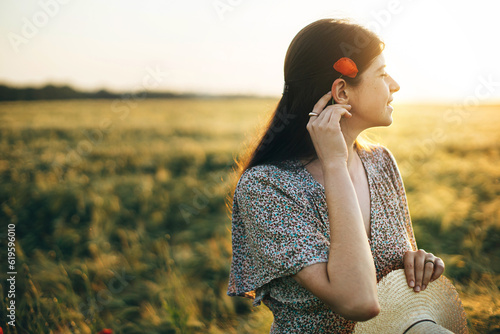 Beautiful woman with poppy flower behind ear relaxing in barley field in sunset light. Stylish female standing in evening summer countryside. Atmospheric moment, rustic slow life