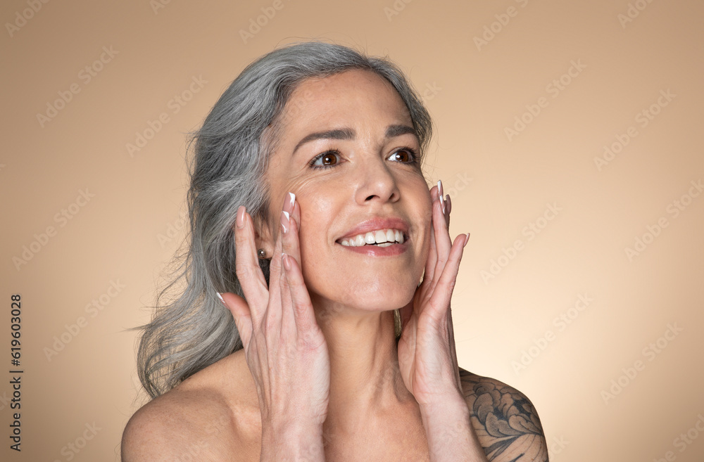 Beautiful senior woman touching her smooth face skin, applying moisturizer cream or lotion on cheeks, beige background Beautiful senior woman touching her smooth face skin, applying moisturizer cream or lotion on cheeks, beige background
