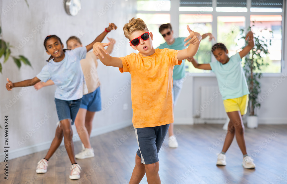 Boy in sun glasses performs choreographic exercises and teaches energetic mobile social dance Jazz-modern together with friends. Multi-racial group of children performs movements in spacious studio