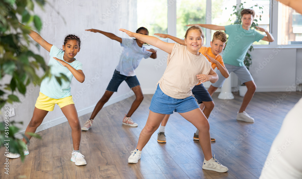 Fototapeta premium Female youngster performs choreographic exercises and teaches energetic mobile social dance hip hop together with friends. Young girls and guys repeat movements, train in spacious studio unfocused