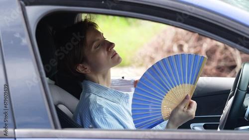 Tired exhausted middle aged woman waving blue fan suffers from stuffiness driving car on summer hot weather. Overheating, sultriness, high temperature, swelter in car with broken air conditioner.