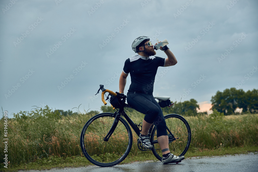 Bearded young man, cyclist in uniform, helmet and glasses standing by the road and drinking water. Cloudy evening. Concept of sport, hobby, leisure activity, training, health, speed, endurance, ad