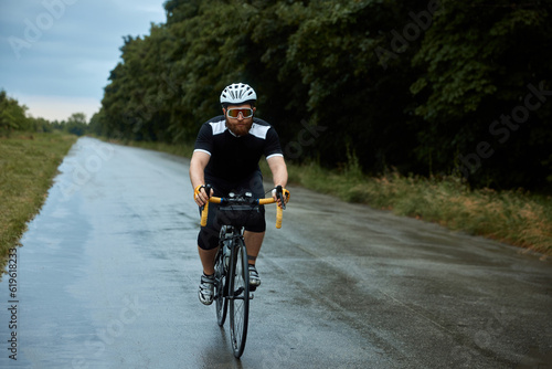 Sportive, young, bearded man, cyclist in uniform, helmet and glasses riding bike on wet road in chill evening after rain. Sport, hobby, leisure activity, training, health, speed, endurance, ad concept © master1305