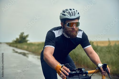 Young bearded man, cyclist in helmet, glasses and uniform riding bike on wet road in cloudy chill evening. Concept of sport, hobby, leisure activity, training, health, speed, endurance, ad