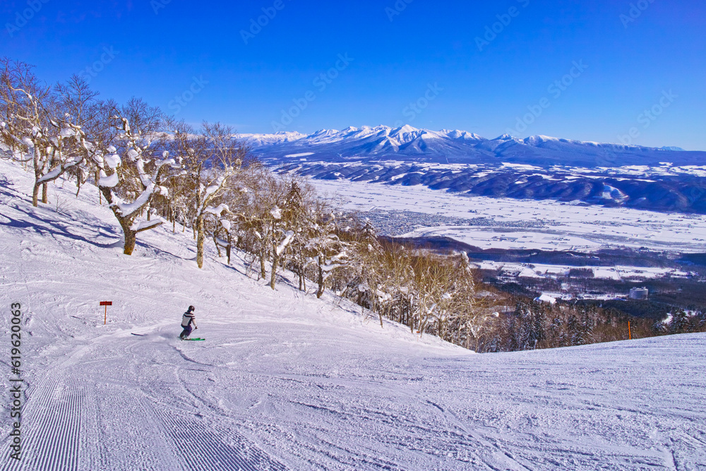 快晴の富良野スキー場から見る十勝岳連峰、富良野ゾーンの標高最高地点からの中級者コースを滑るスキーヤー
