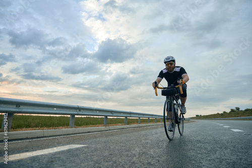 Dynamic image of young bearded sportive man, cyclist in helmet riding bike on empty road in the evening. Concept of sport, hobby, leisure activity, training, health, speed, endurance, ad