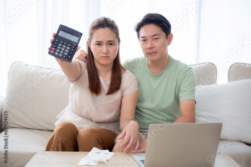 Unhappy young asian couple sitting on sofa show calculator with loan and expense in living room at home, man and woman planning about financial and mortgage, lifestyles and finance concept.