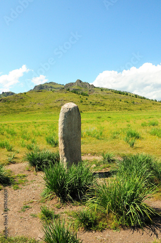 A tall white granite monolith surrounded by low bushes stands in a picturesque valley surrounded by high mountains.