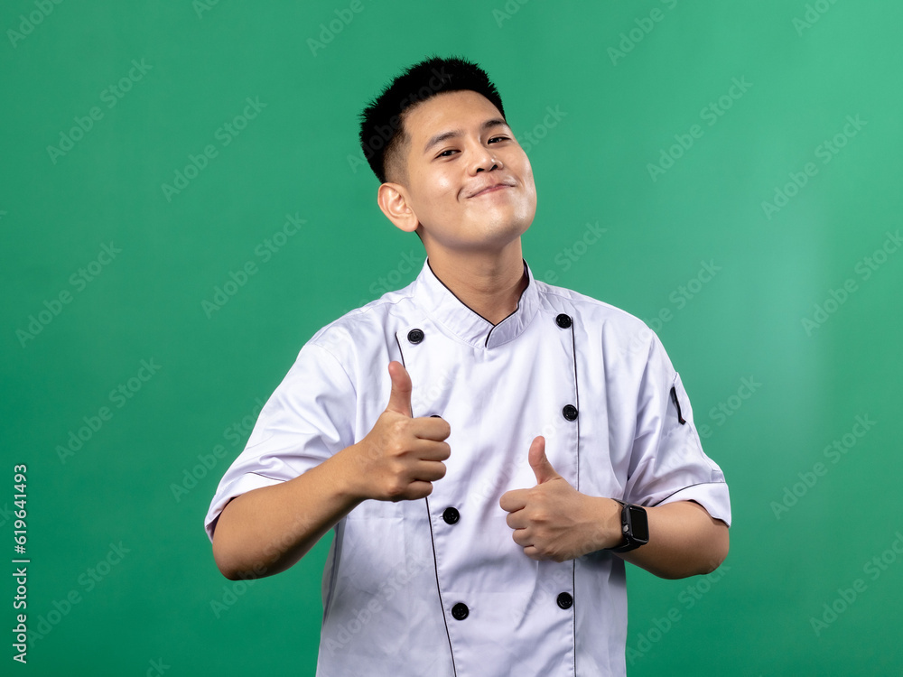 A portrait of an Indonesian Asian man wearing a chef's uniform and a chef's hat, posing with a thumbs up gesture, isolated with a green background. A portrait of an Indonesian Asian man wearing a chef's uniform and a chef's hat, posing with a thumbs up gesture, isolated with a green background.