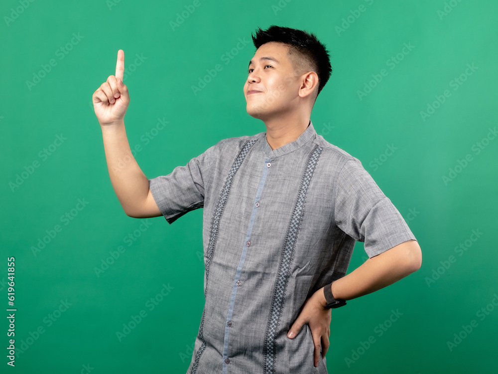 A portrait of an Indonesian Asian man wearing a gray shirt, looking happy and pointing upwards with one hand while the other hand is on his hip, isolated against a green background.