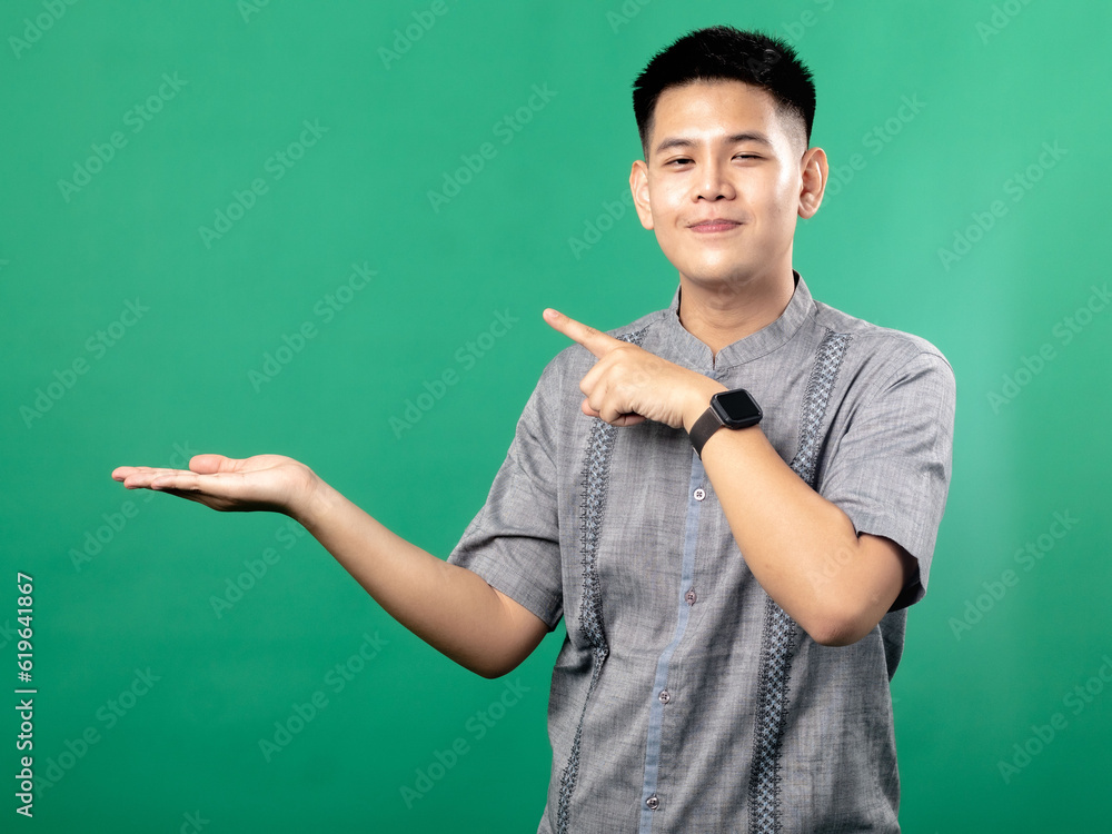 A portrait of an Indonesian Asian man wearing a gray shirt, looking happy and posing with one hand holding an empty item while the other hand points towards it, isolated against a green background.