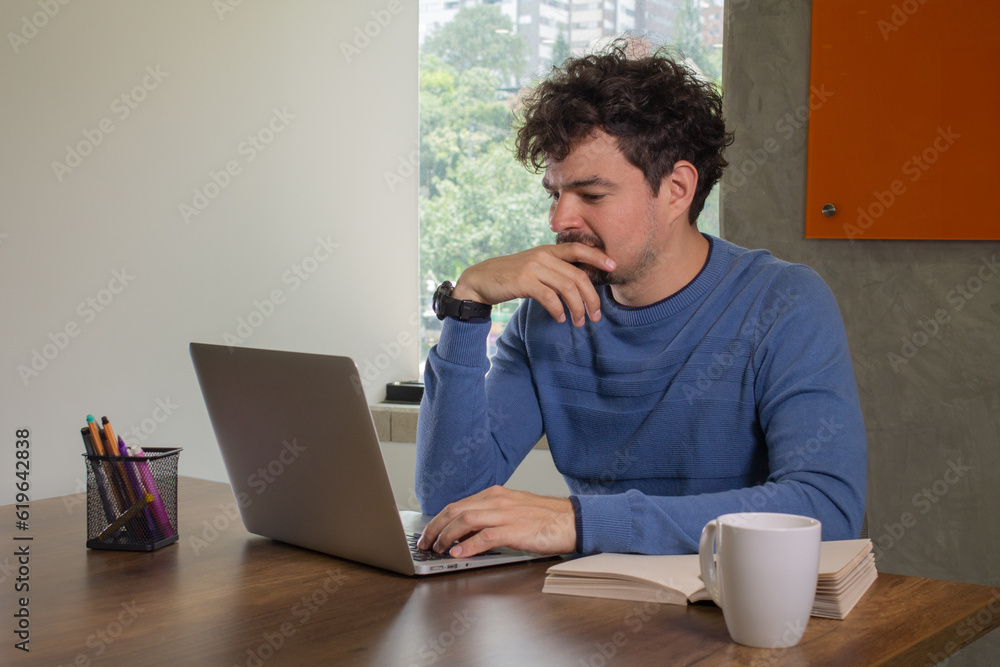 A tired and worried young Latin man is working on his laptop in a casual office, fully focused on his work. A tired and worried young Latin man is working on his laptop in a casual office, fully focused on his work.