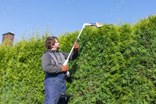 Male gardener using a long reach pole hedge trimmer to cut the top of a tall hedge. Professional gardener with a professional garden tools at work.