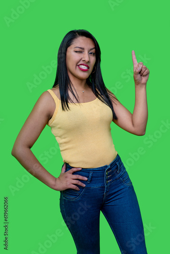 overjoyed brunette woman with one eye winking in yellow tank top pointing at something, video art, archival photo, studio portrait, studio photography green background