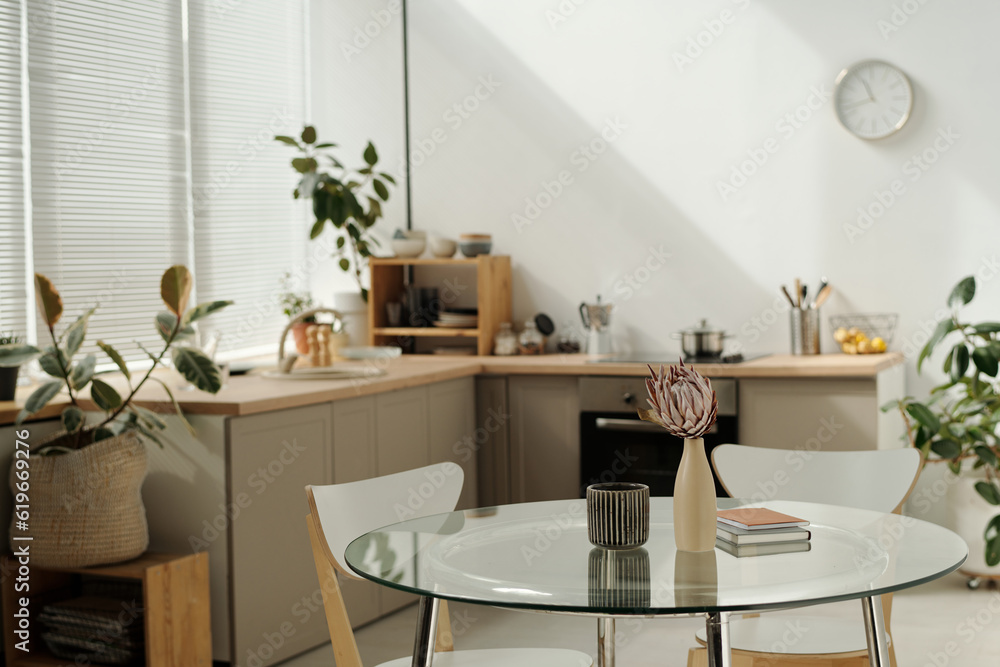 Part of spacious kitchen with round glass table with vases and notepads standing in the center against green domestic plants and counter Part of spacious kitchen with round glass table with vases and notepads standing in the center against green domestic plants and counter