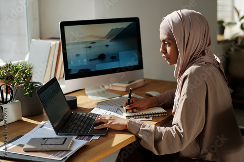 Young serious Muslim businesswoman looking at laptop screen and making notes in notepad while analyzing online data by workplace