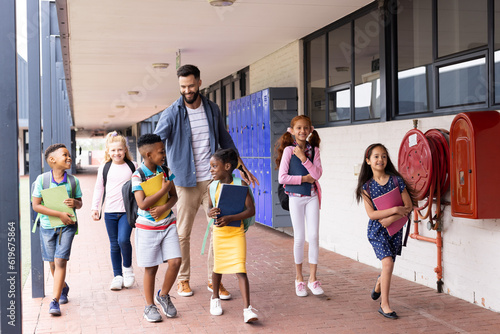Diverse  happy male teacher and elementary schoolchildren walking in school corridor  copy space