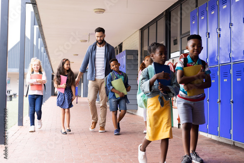 Diverse, happy male teacher and children walking together in elementary school corridor, copy space