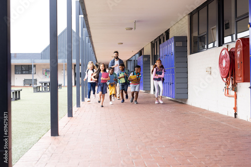 Diverse male teacher with happy children running in elementary school corridor, copy space