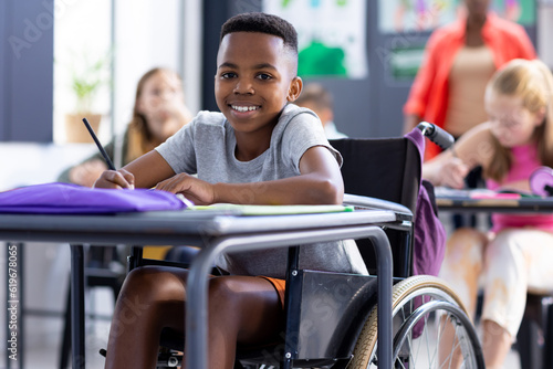 Portrait of smiling african american schoolboy in wheelchair working at desk in class photo