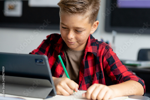 Happy caucasian schoolboy sitting at desk using tablet in class and writing