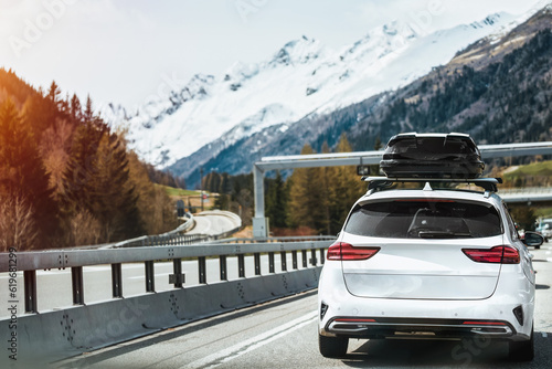 Black Roof Box on a Sporty White SUV Family Car. Rooftop cargo carrier bag. Rear view of a car with a roof box. Alpine highway. Removable black car trunk for luggage on the roof of a car. © AlexGo