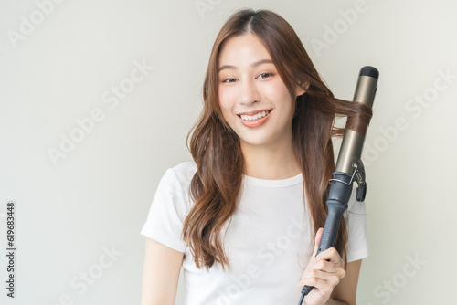 Fototapeta Beautiful curly brunette, attractive asian young woman, girl smiling using curler, curling iron long hair with ceramic curler straightener at home