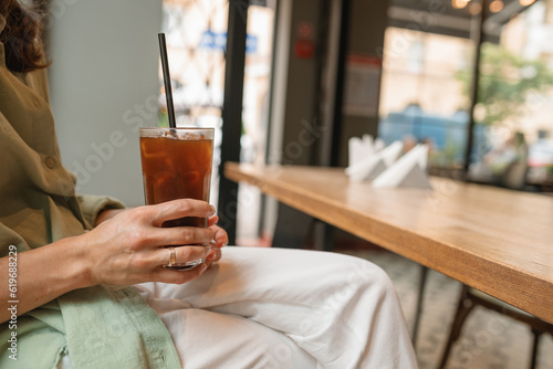 Close-up of woman in green linen shirt and white trousers sitting in cafe and drinking espresso tonic.