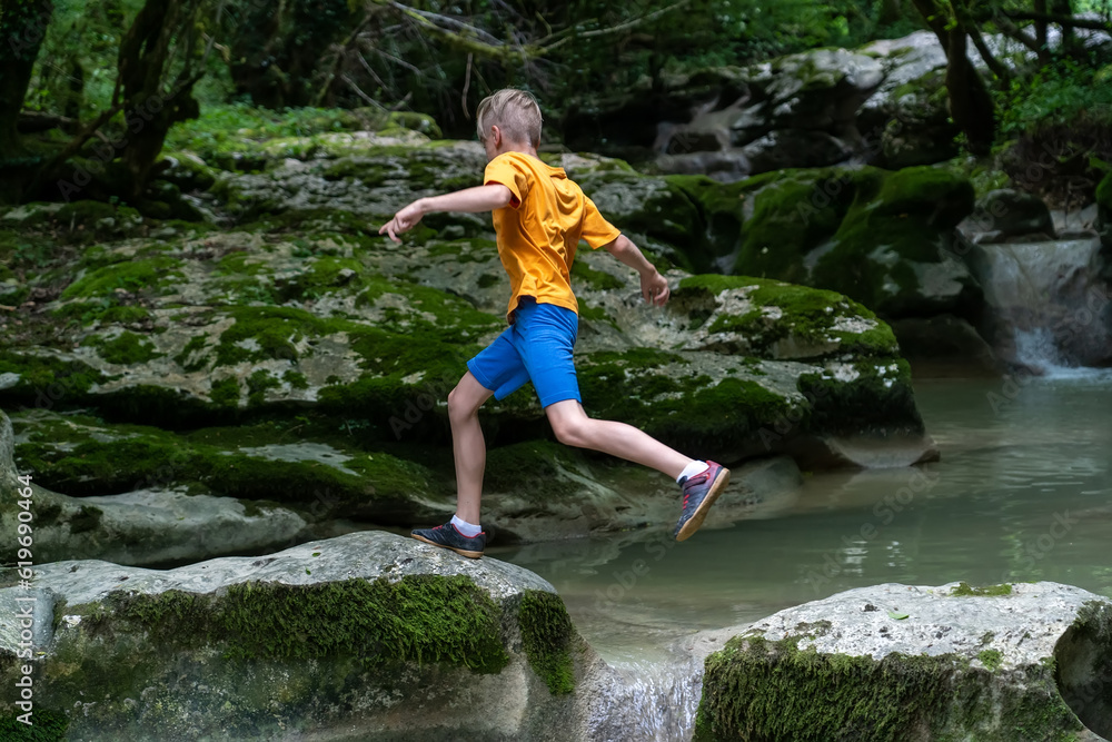 Active boy jumps from rock to rock on a calm mountain river hidden in a dense forest Active boy jumps from rock to rock on a calm mountain river hidden in a dense forest