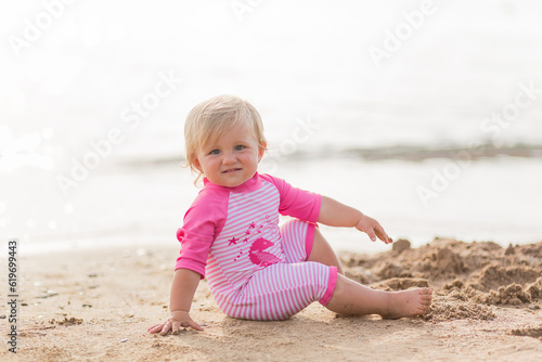 a cute little happy smiling tanned caucasian kid toddler baby girl in one-piece striped pink swimsuit plays in the sand  on the beach. Water safety. Vacation and family holiday 