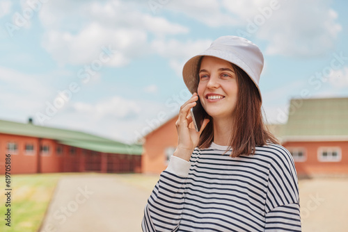 Attractive satisfied joyful dark haired woman wearing striped shirt and panama walking on streets talking mobile phone smiling while looking away.
