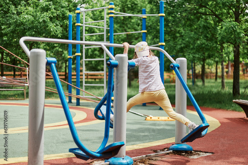 Full length portrait of little girl wearing baseball cap doing physical exercises on sportsground dressed casual closing making training on legs on on outdoor simulator. photo