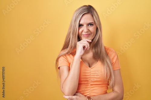 Young woman standing over yellow background looking confident at the camera smiling with crossed arms and hand raised on chin. thinking positive.
