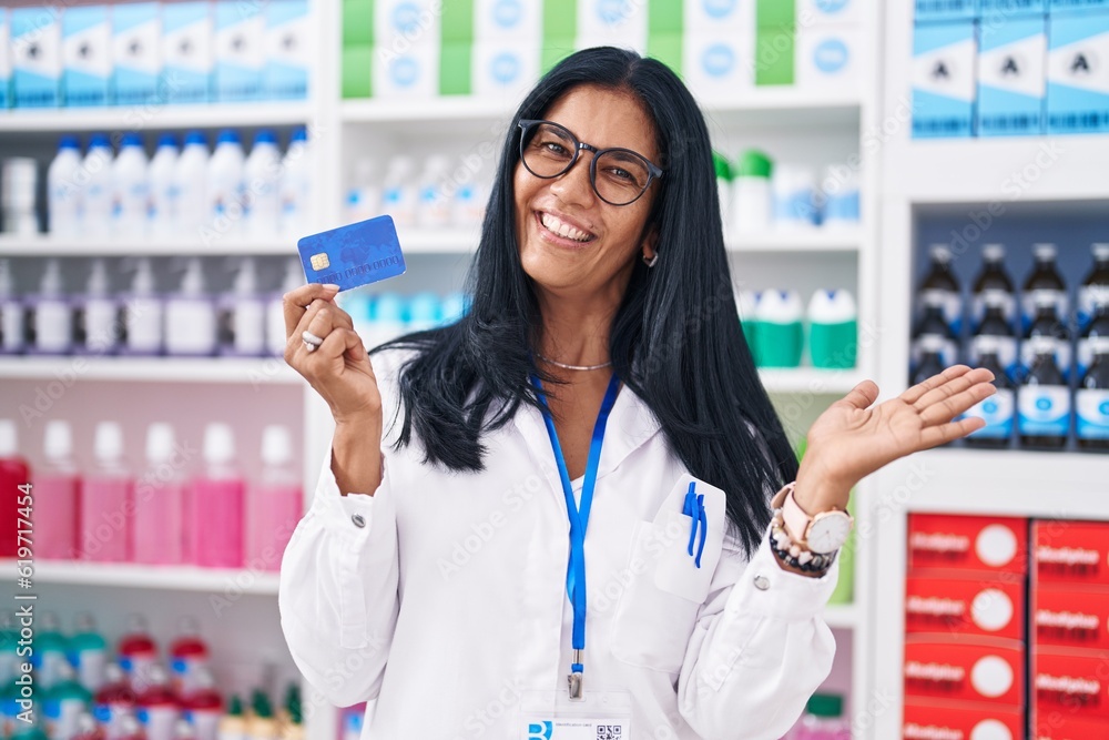 Mature hispanic woman working at pharmacy drugstore holding credit card celebrating achievement with happy smile and winner expression with raised hand
