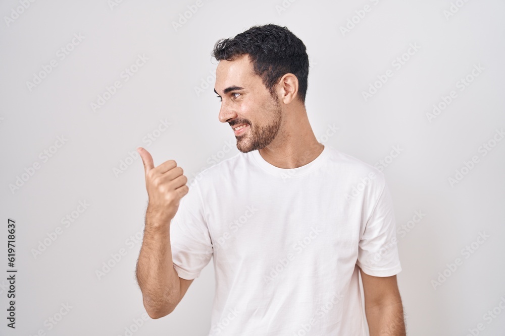 Handsome hispanic man standing over white background smiling with happy face looking and pointing to the side with thumb up. Handsome hispanic man standing over white background smiling with happy face looking and pointing to the side with thumb up.