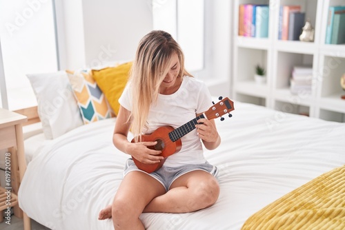 Young blonde woman playing ukulele sitting on bed at bedroom