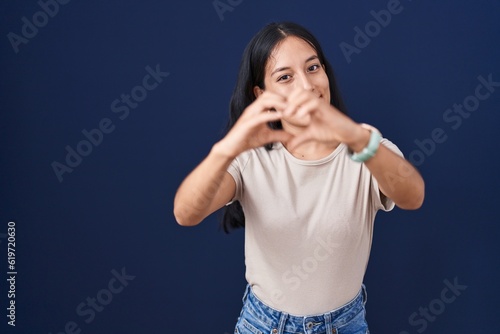 Young hispanic woman standing over blue background smiling in love doing heart symbol shape with hands. romantic concept.