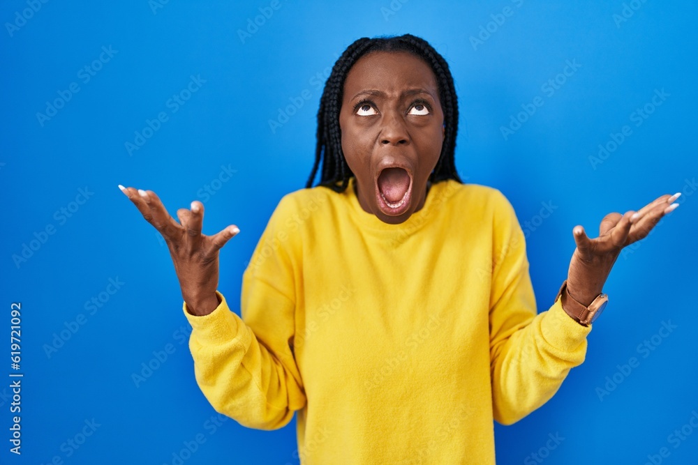 Beautiful black woman standing over blue background crazy and mad shouting and yelling with aggressive expression and arms raised. frustration concept. Beautiful black woman standing over blue background crazy and mad shouting and yelling with aggressive expression and arms raised. frustration concept.
