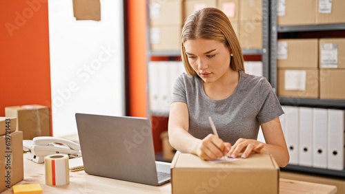 Young blonde woman ecommerce business worker using laptop writing on package at office