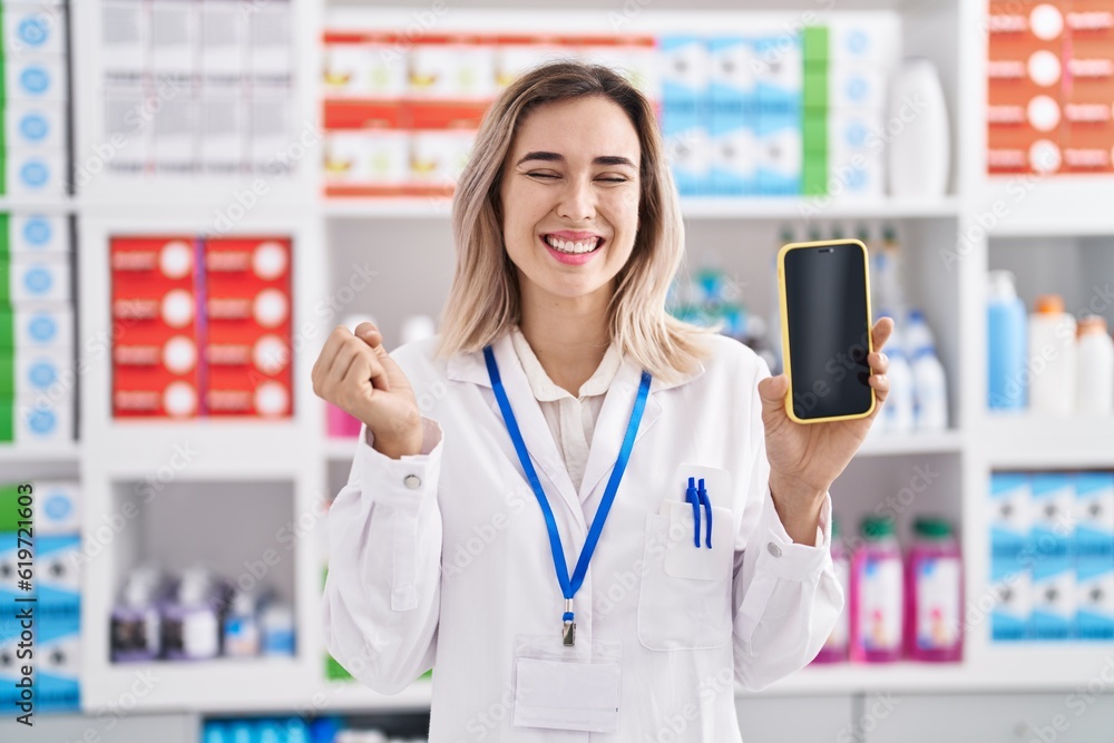 Young beautiful woman working at pharmacy drugstore showing smartphone screen screaming proud, celebrating victory and success very excited with raised arm