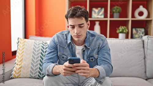 Young hispanic man using smartphone sitting on sofa at home