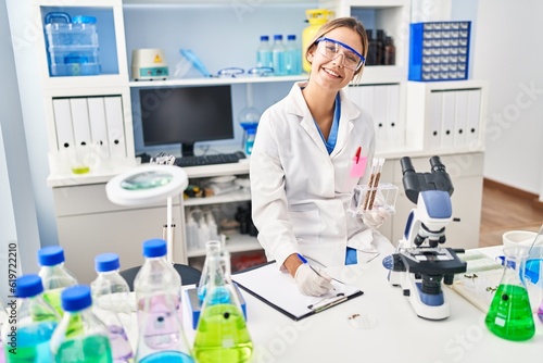 Young hispanic woman scientist write on document holding test tubes at laboratory