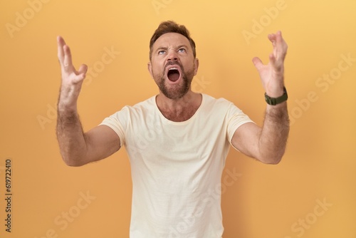 Middle age man with beard standing over yellow background crazy and mad shouting and yelling with aggressive expression and arms raised. frustration concept.