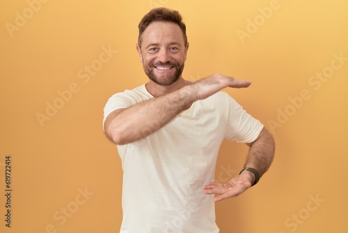 Middle age man with beard standing over yellow background gesturing with hands showing big and large size sign, measure symbol. smiling looking at the camera. measuring concept.