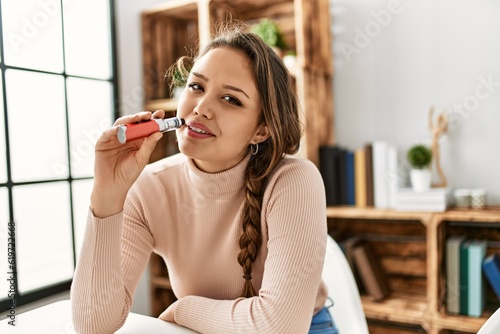 Young beautiful hispanic woman holding vaper sitting on table at home