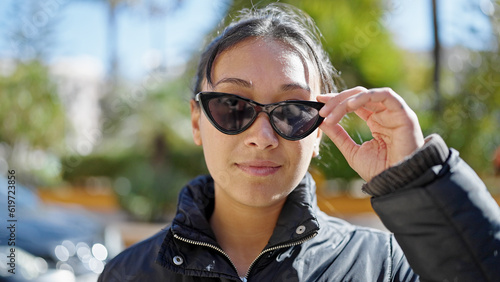 Young beautiful hispanic woman standing with serious expression wearing sunglasses at park
