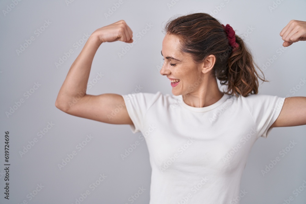 Fototapeta premium Beautiful brunette woman standing over isolated background showing arms muscles smiling proud. fitness concept.