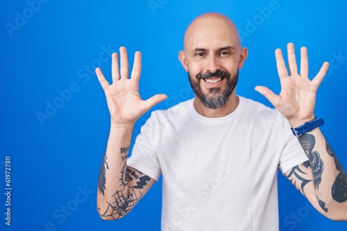 Hispanic man with tattoos standing over blue background showing and pointing up with fingers number ten while smiling confident and happy.