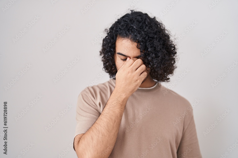 Hispanic man with curly hair standing over white background tired rubbing nose and eyes feeling fatigue and headache. stress and frustration concept. Hispanic man with curly hair standing over white background tired rubbing nose and eyes feeling fatigue and headache. stress and frustration concept.