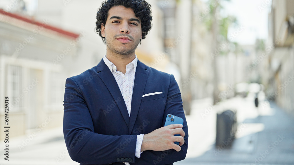 Young latin man business worker standing with arms crossed gesture holding smartphone at street Young latin man business worker standing with arms crossed gesture holding smartphone at street